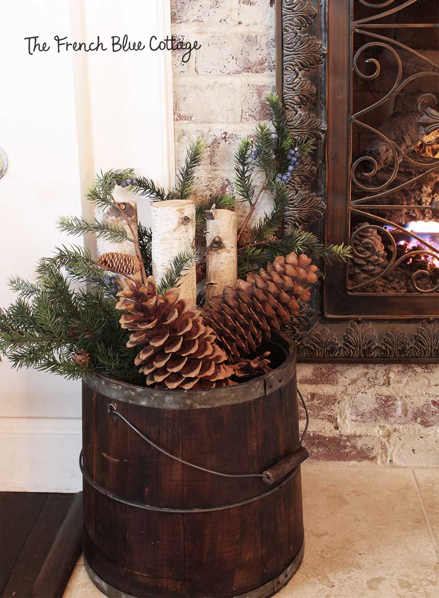 Birch logs, evergreens, and pinecones in large bucket by fireplace.