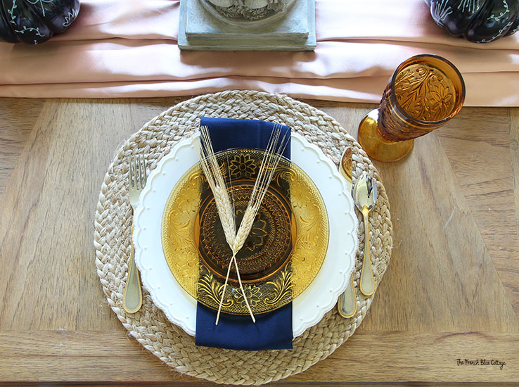 fall place setting with a round jute placemat, white plate, navy napkin, and amber salad plate and glass. Two sprigs of wheat are on the plate as decoration.