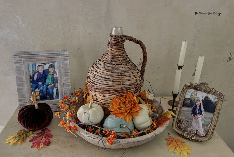 fall vignette with a wicker bottle in a wooden bowl surrounded by flowers and small pumpkins