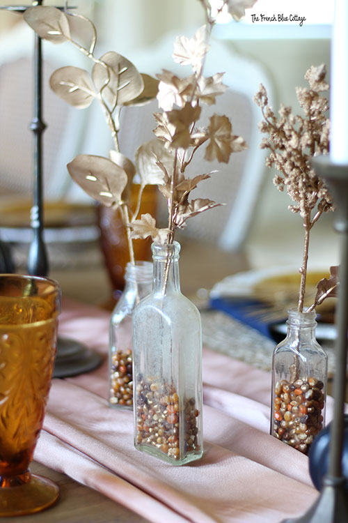 brown, gold, and copper spray painted corn kernels is the vase filler in these clear bottles with a single gold stem of leaves