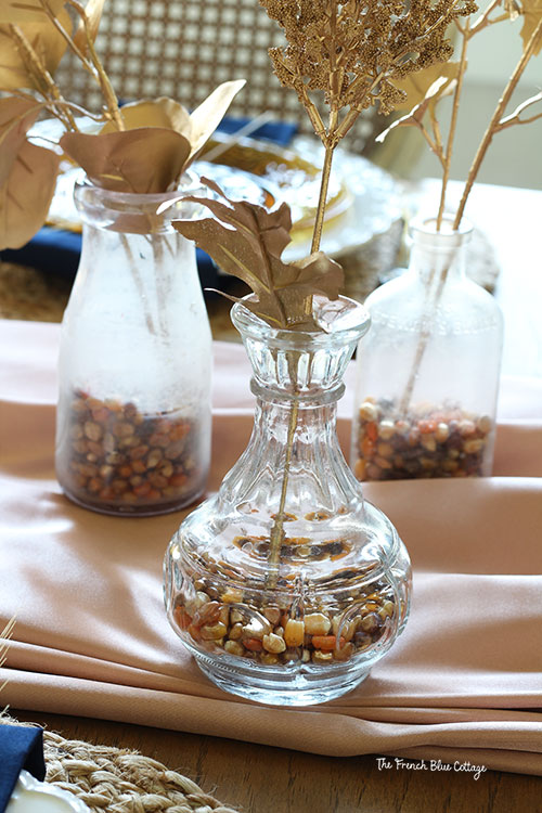 brown, gold, and copper spray painted corn kernels is the vase filler in these clear bottles with a single gold stem of leaves